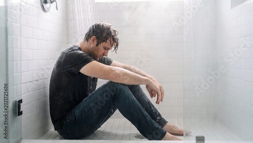 A man sits alone and dejected on the cold floor of a modern shower, head bowed in quiet despair, reflecting on personal struggles amidst the steamy bathroom