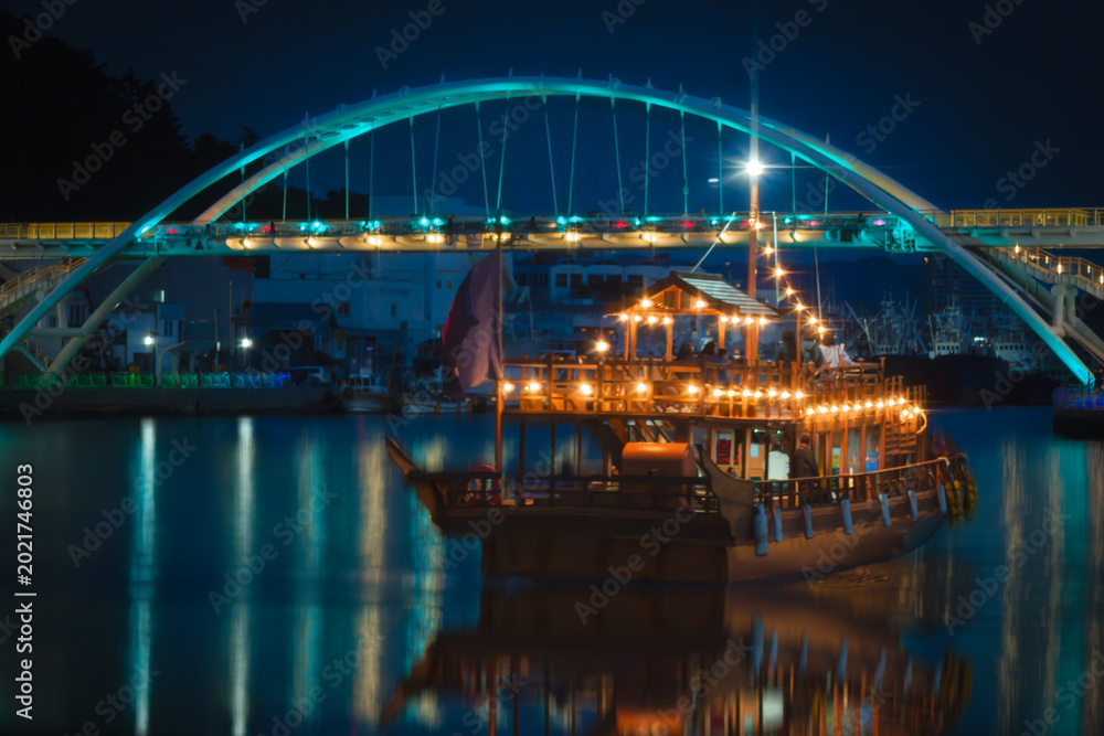 Fototapeta premium Motion blur night Seascape at the illuminated Gangguan Port with night cruise passing the arching bridge in Tongyeong‑si, Gyeongsangnam‑do, South Korea