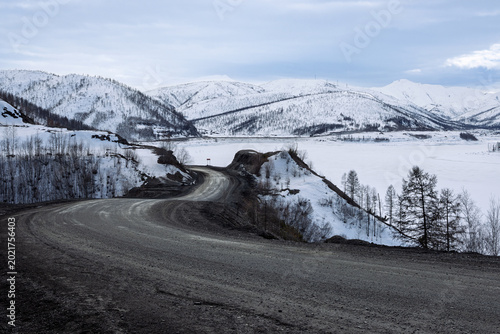 A landscape with a winding road through the mountains. The ice-covered Ust-Srednekanskoye Reservoir is visible on the right. Snow-capped hills are visible in the distance. Magadan Region, Russia.
