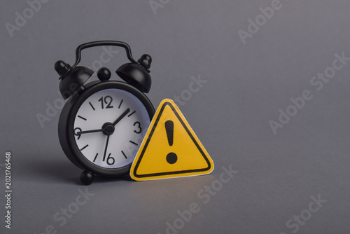 Alarm clock with warning symbol on gray background representing deadline alerts, urgency, risk awareness, time pressure, reminder, and business warning concepts.