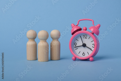 Pink alarm clock beside wooden human figures on blue background representing teamwork, scheduling, punctuality, productivity, meeting management, and business organization concepts.