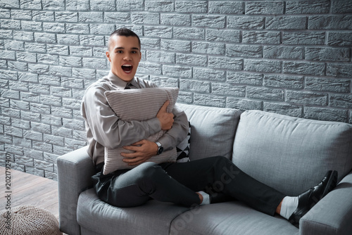 Foto Afraid young man screaming and sitting on sofa in living room near brick wall