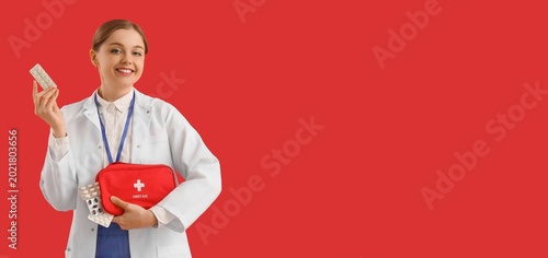 Female doctor holding first aid kit and pills on red background with space fo...