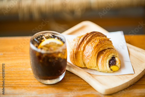 Foto Croissant and Refreshing Beverage: A tempting assortment featuring a golden, flaky croissant alongside a chilled glass of invigorating cola, both artfully presented on a wooden tray