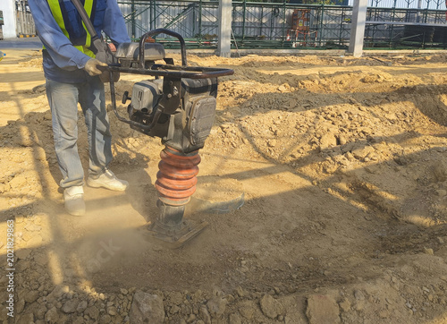 Εκτύπωση καμβά A construction worker is using a jumping jack compactor to compact soil in a factory area for construction site preparation
