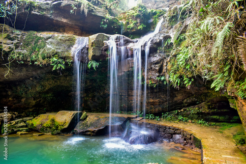 Fotografija Scenic tropical waterfall cascading over moss-covered rocks into a crystal-clear pool, surrounded by lush green forest vegetation and serene natural landscape in The Garden of Caves, Meghalaya