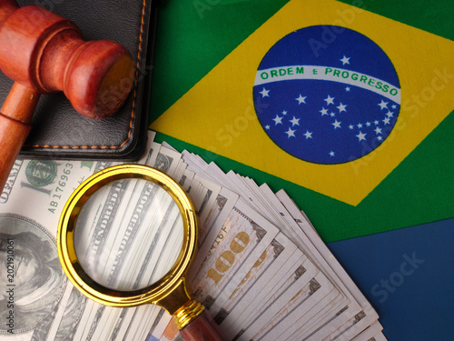 A wooden gavel, magnifying glass, and stack of dollar bills are placed over a Brazilian national flag