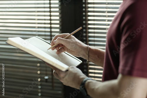 Caucasian adult man writing notes in notebook while standing indoors, capturing planning and focus
