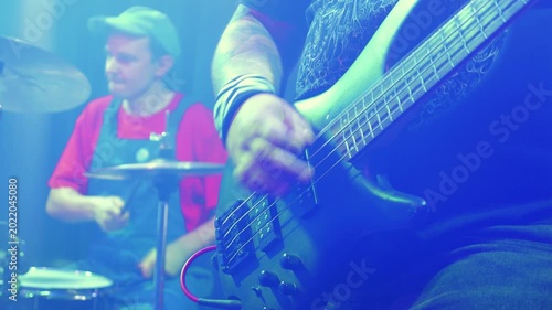 Close up of tattooed bassist playing bass guitar. Drummer in background at live concert. Energetic punk rock show with vibrant blue stage lighting and smoky atmosphere