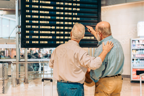 Rear view of senior travelers checking flight information board together at airport terminal