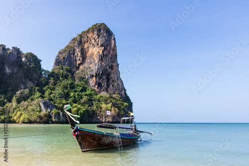 Longtail boat and tall limestone cliff, on the tropical shores of Railay Beach in Krabi, southern Thailand. Andaman Sea on the background. 
