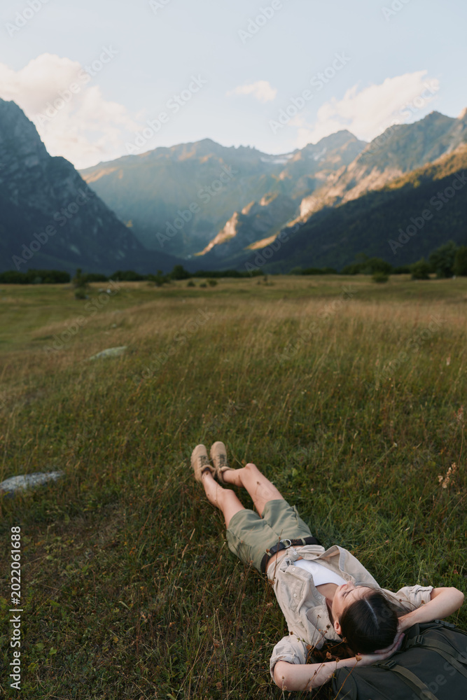 Fototapeta premium Woman relaxing in a grassy field near mountains, enjoying outdoor life, casual outfit, lying on her back with arms behind her head, serene natural scenery and peaceful mood