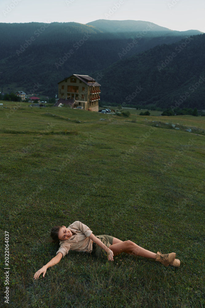 Fototapeta premium A woman lies on a grassy field outdoors, distant house and mountains in the background, conveying relaxation, leisure, and a moment of nature inspired calm.