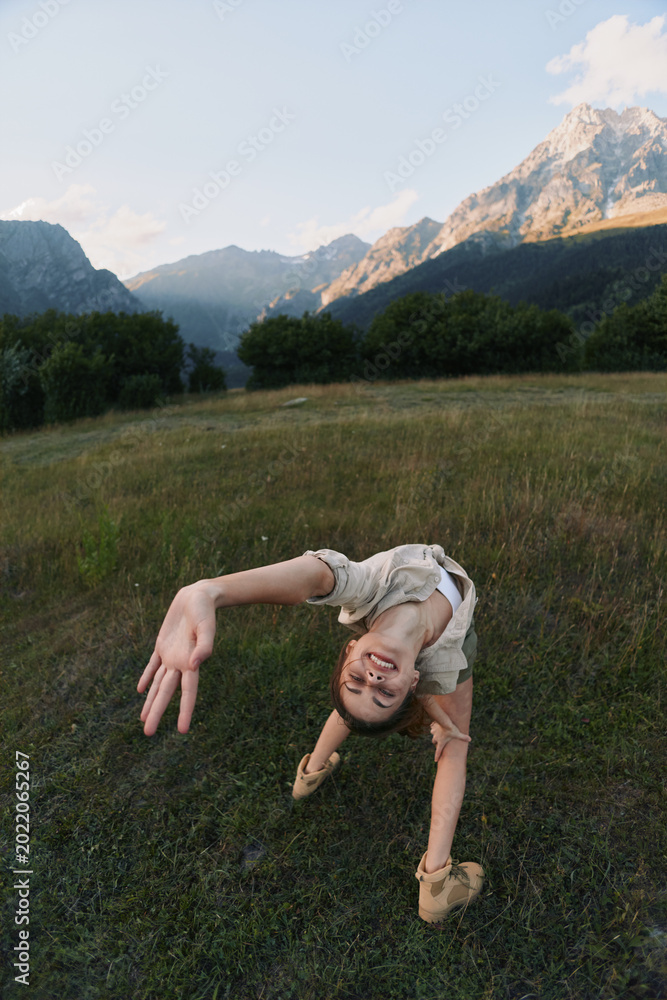 Fototapeta premium Young woman in a meadow outdoor scene performing a backbend, smiling at the camera, wind in hair, mountains in the background, colorful nature, relaxed lifestyle moment