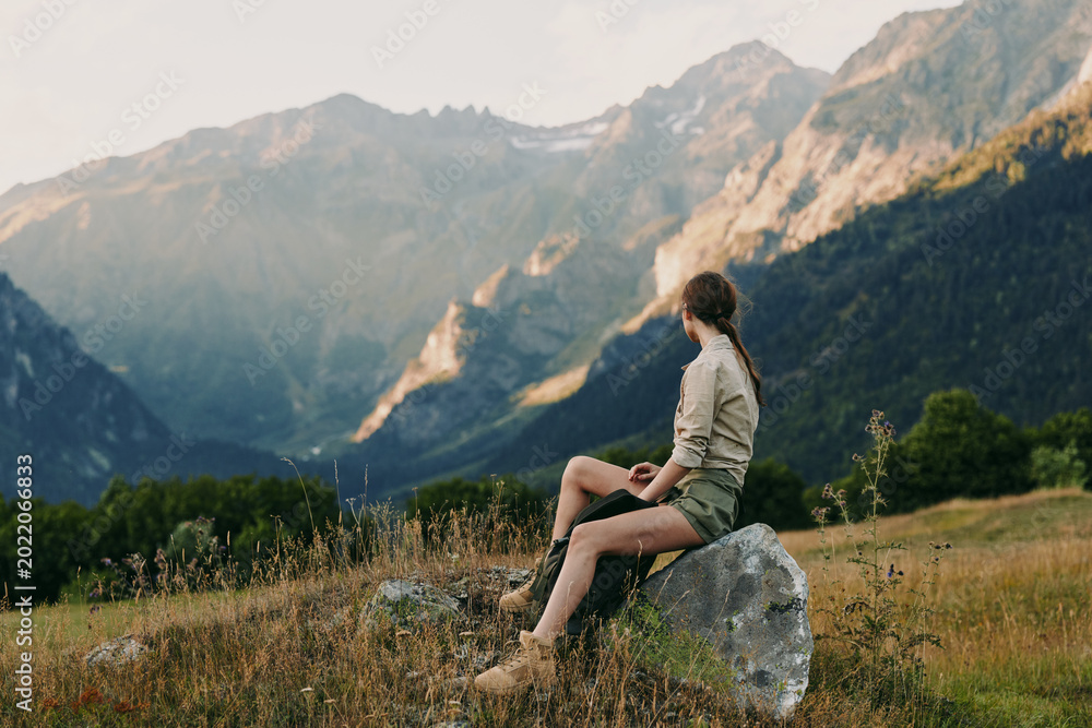 Fototapeta premium Woman sits on a rock in a mountain meadow, enjoying vast valleys and blue skies. Casual outdoor lifestyle shot emphasizing nature, wellness, and solitude.