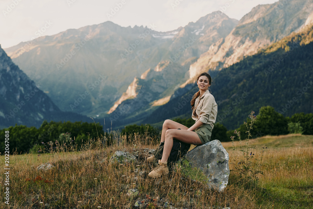 Fototapeta premium Young woman lifestyle sitting on rock in mountain meadow, casual outfit, relaxed pose, natural light, scenic landscape background, warm atmosphere