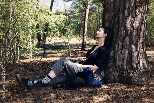 Young woman rests against large pine tree trunk in sunlit forest with eyes closed. Mental health awareness, emotional balance, inner peace concept, Stress relief nature.