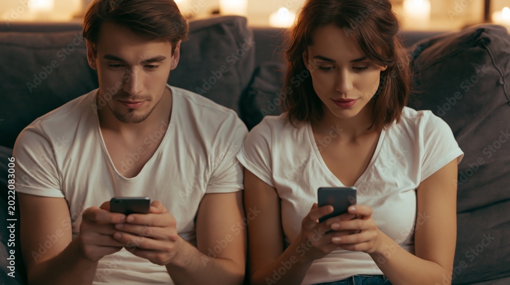 Fototapeta premium Couple Sits Together but Focuses on Smartphones Instead of Each Other in a Living Room Setting With Low Light