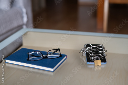 Smartphone wrapped in a metal chain and secured with a padlock, resting on a beige surface next to a blue book and reading glasses, representing digital detox, mindful living, and online security