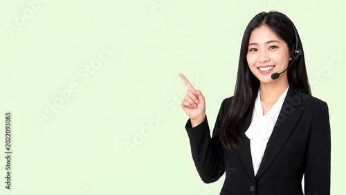 Smiling Asian female customer support agent wearing a headset and pointing finger at copy space. Professional customer care representative in business suit showing blank presentation space.