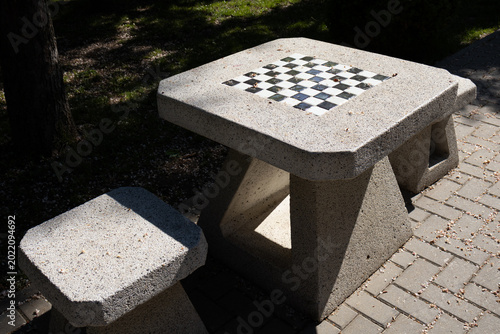 A complete outdoor gaming station featuring a stone table with a chessboard and matching concrete stools in a sunny park corner
