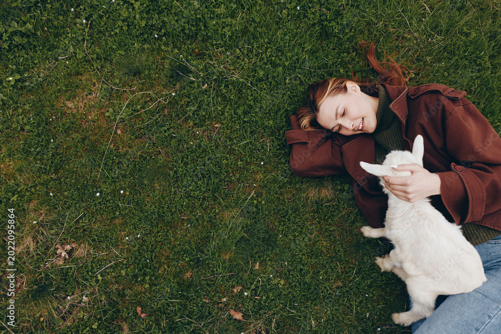 Fototapeta premium Cheerful outdoor moment of a woman lying on grass, cuddling a white goat kid, warm sunlight, relaxed posture, soft breeze, natural colors, candid lifestyle scene, gentle affection