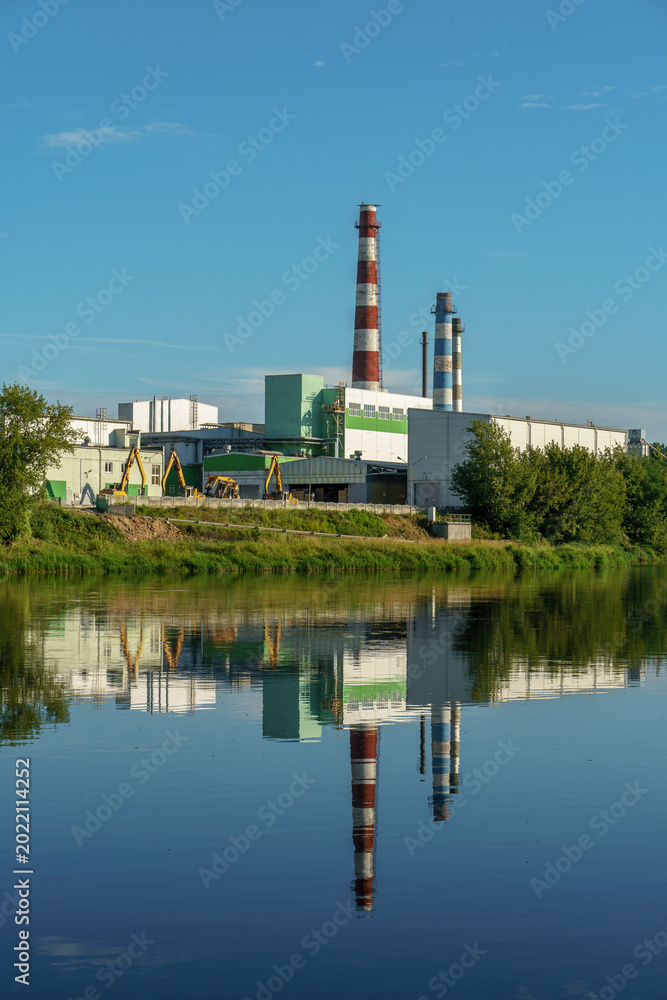 Fototapeta premium An industrial factory on the riverbank. The factory's reflection in the water. Smoke from the chimney of a furniture factory