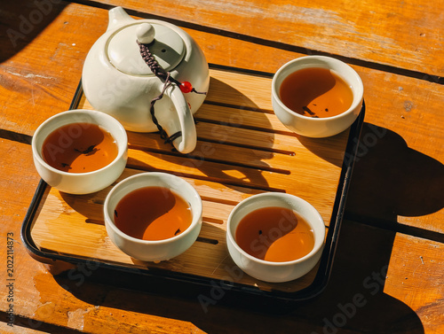 Chinese tea set with white teapot and four cups on bamboo tray. A white ceramic teapot and four cups filled with amber tea on bamboo tray. Bright sunlight creates sharp shadows on rustic wooden table.