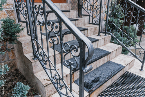 Stone staircase steps with black anti slip mats and ornate dark gray wrought iron handrail. Architecture design detail