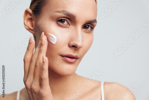 Woman applying white facial cream on her cheek. Close up portrait with soft lighting and a neutral background. Skincare, wellness, and self care themes emphasized in a calm, clean mood.