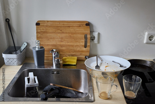 Fotografi A messy kitchen sink counter filled with dirty dishes, unwashed glass mugs, greasy plates with cutlery, and a cutting board