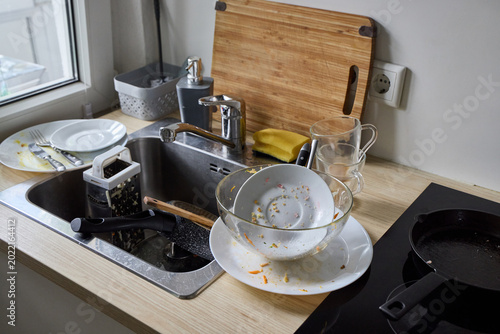 Fotografía A messy kitchen sink counter filled with dirty dishes, unwashed glass mugs, greasy plates with cutlery, and a cutting board