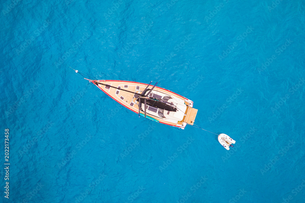 Fototapeta premium High-angle aerial drone shot of a red and white sailboat anchored in crystal-clear turquoise ocean water. A serene image capturing the essence of a luxury summer sailing holiday and tropical adventure