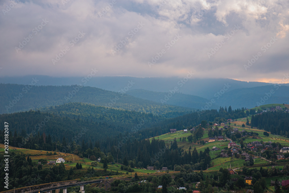 Fototapeta premium Picturesque mountain landscape. Ukrainian Carpathians. A cozy village lies in the valley below, surrounded by rolling green hills and misty mountain peaks. Bright traditional houses and untouched