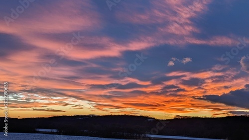 Dramatic sunset landscape with glowing orange and pink clouds above dark hills and winter fields, creating a vivid evening sky.