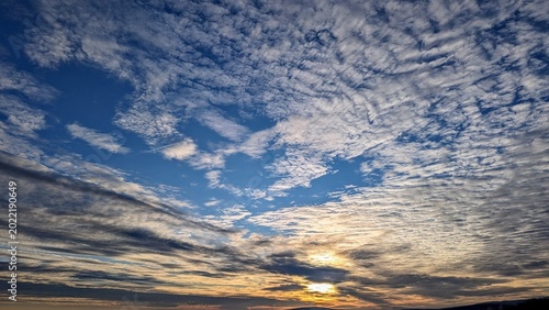 Wide sunset view with dramatic textured clouds, golden sunlight, blue sky, and dark silhouetted hills on the horizon.