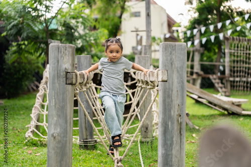 preschool child girl playing and walking on wooden rope bridge at outdoor playground in park
