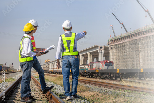 Group of engineers discussing infrastructure project at a railway construction site, industrial foreman and workers in safety gear looking at bridge construction near train tracks