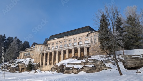 Historic stone mountain building surrounded by snow, rocky terrain, frosty trees, and clear blue sky in a winter landscape, photographed in a scenic alpine resort area.