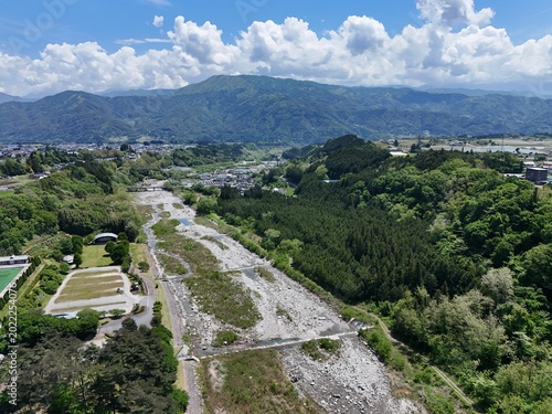 Aerial view of a dry riverbed winding through lush green hills. Clouds drift above distant mountain ridges. Vibrant foliage surrounds the exposed earth. Nature's quiet resilience dominates the scene