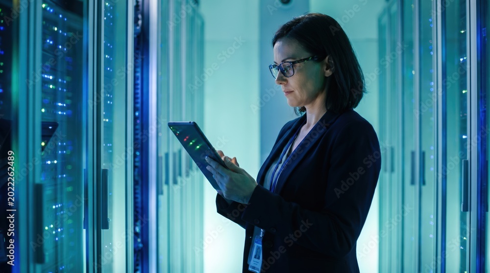 Fototapeta premium Female IT engineer monitoring server racks with digital tablet in data center. Cloud computing and network management.