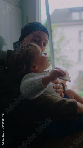 Mother undergoing chemotherapy holding toddler by window at home, headscarf parent supporting child on lap, caregiving moment, maternal bonding, resilience and quiet family connection