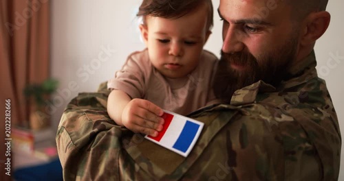 Male french soldier in military uniform hugs his little son at home while baby boy touching french flag - Emotional military happy homecoming concept