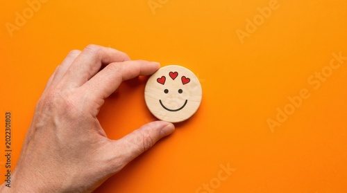 A hand holding a small wooden smiley face with hearts on an orange background