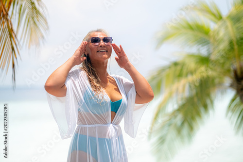 Картината върху платно Asian smiling woman in a blue bikini enjoys a Maldives vacation, with blonde hair and sunglasses, overlooking a palm-tree lined beach