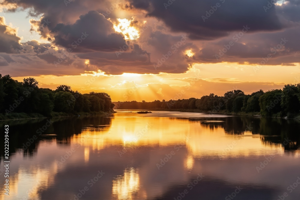 Obraz premium Golden sun rays break through dark clouds at sunset reflecting on calm river water. Lush green forest lines riverbanks. Peaceful summer evening.