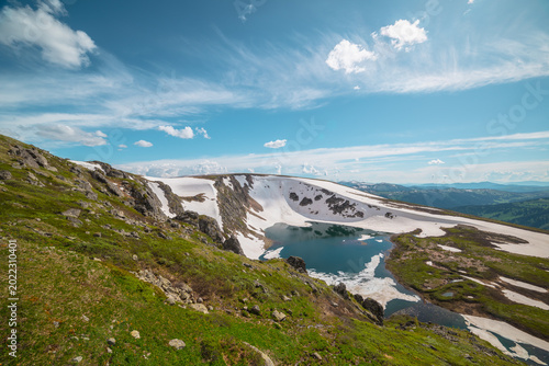Scenic sunlit landscape with alpine lake in rocky snowy cirque near stone hil...