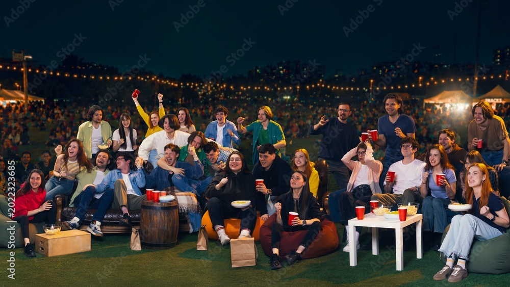 Obraz premium Young people cheering at outdoor night watch party with drinks and snacks. Concept of live sports viewing, fan celebration, summer festival, social gathering and entertainment ads.