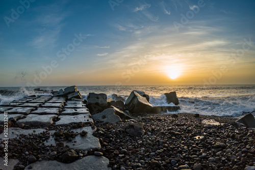 Schilderij op canvas Dramatic sunset over rocky breakwater with powerful waves crashing against stone