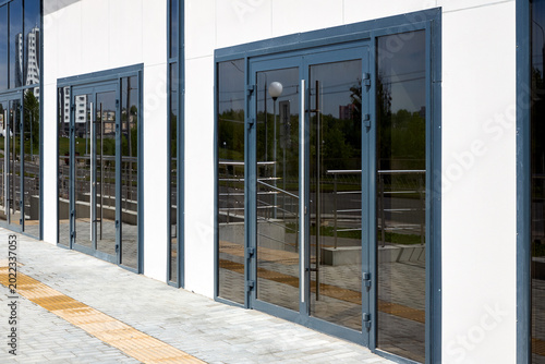 Dark gray aluminum double doors with large glass panels installed in white building facade. Modern architectural design features long vertical handles and matching side windows along paved sidewalk.